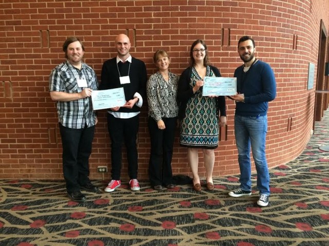 Five people standing in front of a brick wall, holding two large cheques