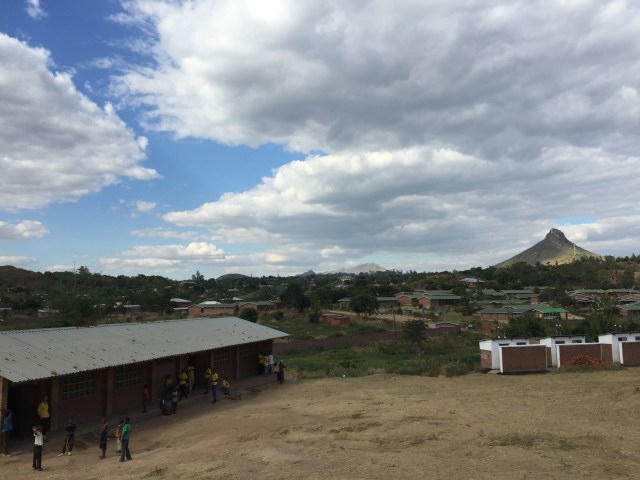 People outside a building with a village, trees and a mountain in the background