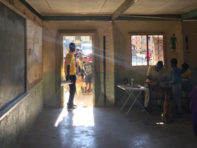 Man in a doorway with a loud hailer, whilst a queue of people wait outside and 2 men at a table process a child and his dog