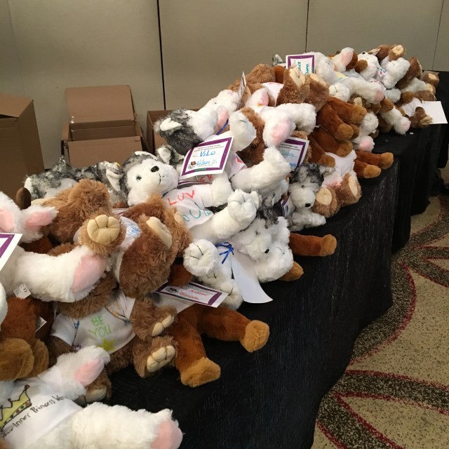 Various teddy bears are piled up on a long table, with open boxes on the floor behind