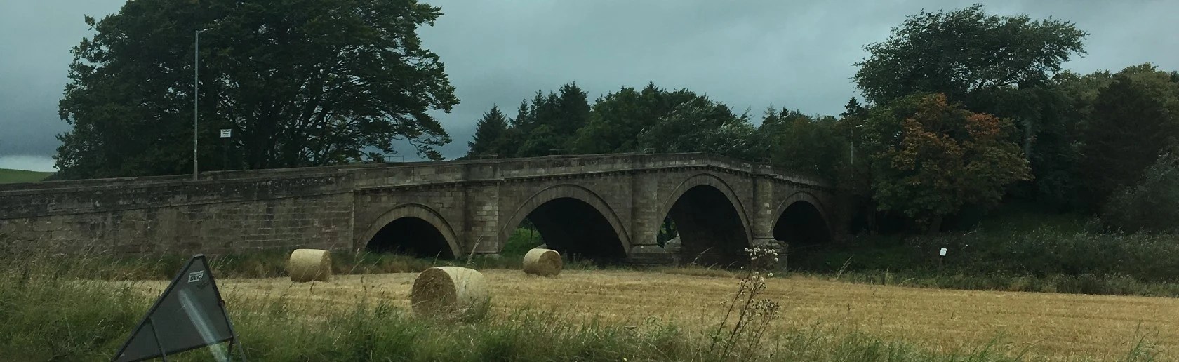 Landscape view of hay bales in a field with an old stone bridge and trees in the distance