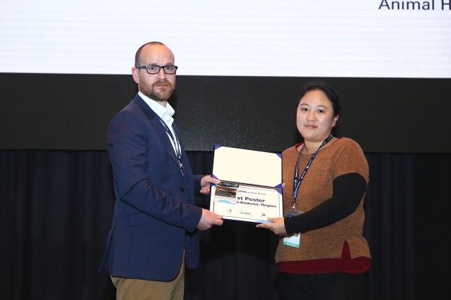 A man is presenting a certificate to a woman, as both look directly into the camera