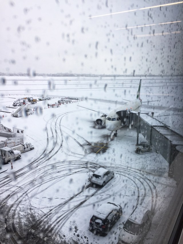 A view from a wet window showing a plane and vehicles on a snowy airport apron