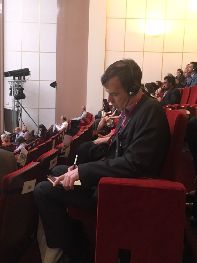 Delegates in a tiered auditorium listening to headsets and writing notes