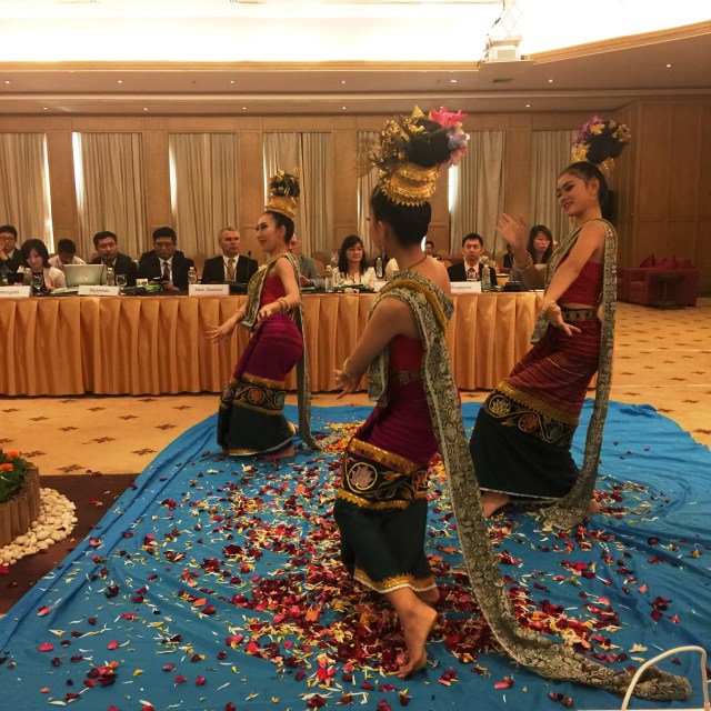 Three Thai women in traditional dress performing for delegates who are sat behind long tables